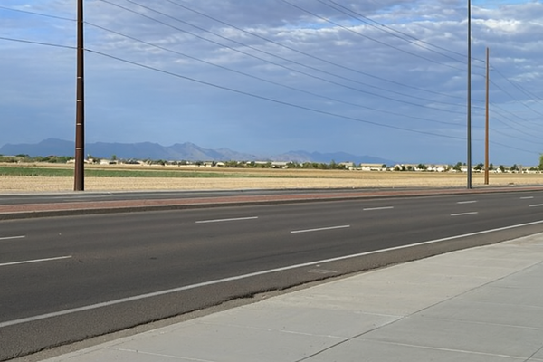 Empty two-lane road in Gilbert, Arizona, under a partly cloudy sky with distant mountains, utility poles, and open land in the background.
