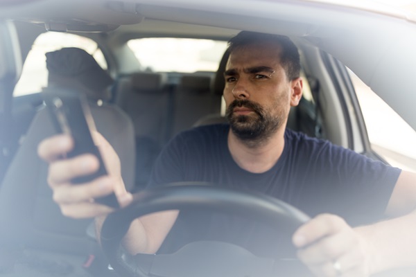 A man with a beard is driving a car while looking down at and holding a smartphone in his left hand, seen through the front windshield.